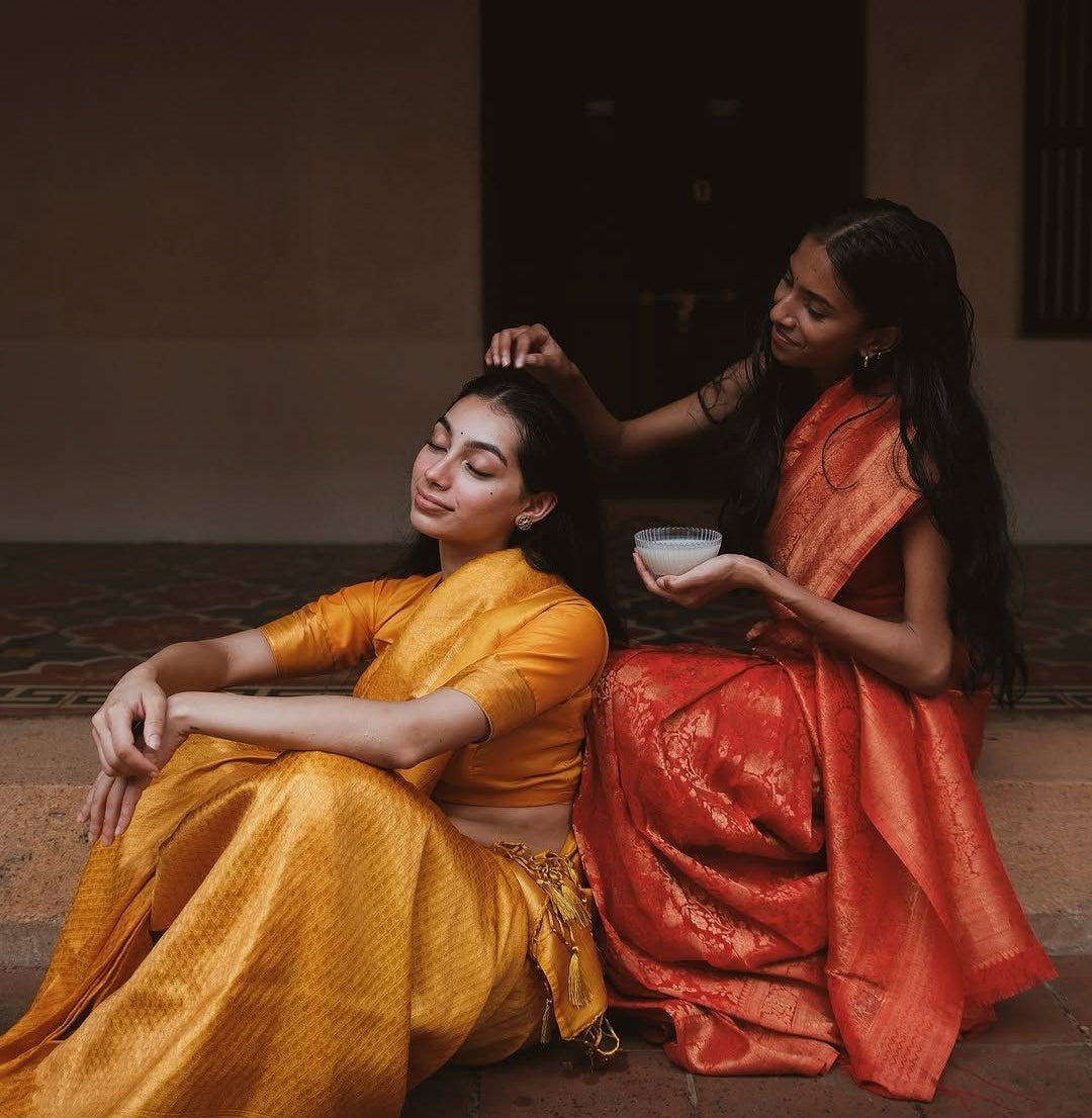 Two women in silk saris, one oiling the other's hair
